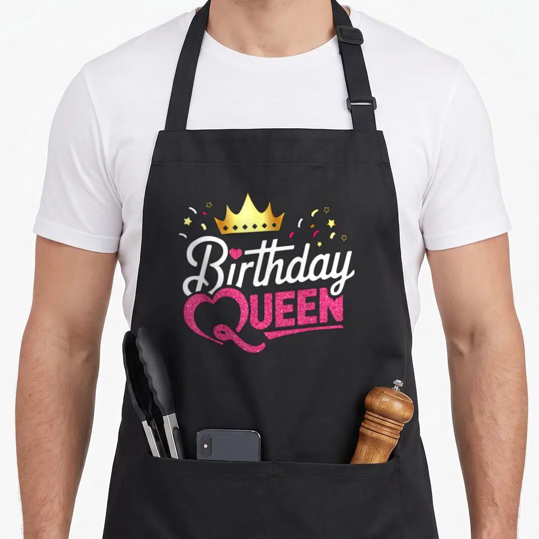 Funny birthday queen of baking apron - Gold crown above white Birthday text and pink QUEEN text with glittery heart — shown on a black cotton canvas apron against a white background