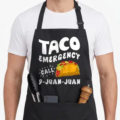 Emergency taco apron - Cartoon taco with text TACO EMERGENCY CALL 9-JUAN-JUAN in white, yellow, red, and green — shown on a black cotton canvas apron against a white background