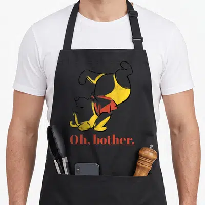 Bother novelty apron - Upside-down yellow bear in red shirt above red Oh bother text — shown on a black cotton canvas apron against a white background