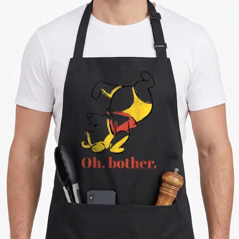 Bother novelty apron - Upside-down yellow bear in red shirt above red Oh bother text — shown on a black cotton canvas apron against a white background