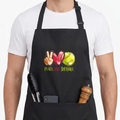 Player i love softball apron - Cartoon peace sign, red heart, and yellow softball with PEACE LOVE SOFTBALL text — shown on a black cotton canvas apron against a white background