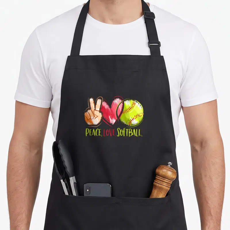 Player i love softball apron - Cartoon peace sign, red heart, and yellow softball with PEACE LOVE SOFTBALL text — shown on a black cotton canvas apron against a white background