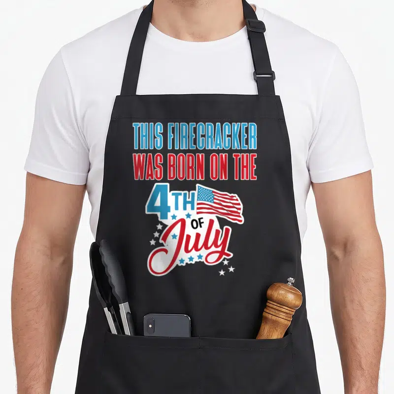 Birthday firecracker 4th of july apron - Stacked red white blue text reading THIS FIRECRACKER WAS BORN ON THE 4TH — shown on a black cotton canvas apron against a white background