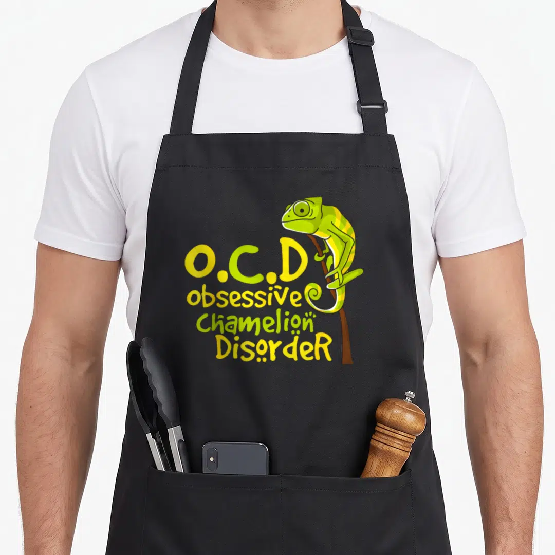 Ocd chameleon lady apron - Cartoon chameleon perched on branch beside stacked yellow and green text reading O.C.D — shown on a black cotton canvas apron against a white background