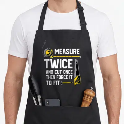 Cut woodworking apron - Stacked text MEASURE TWICE with tape measure saw and hammer in yellow and black — shown on a black cotton canvas apron against a white background