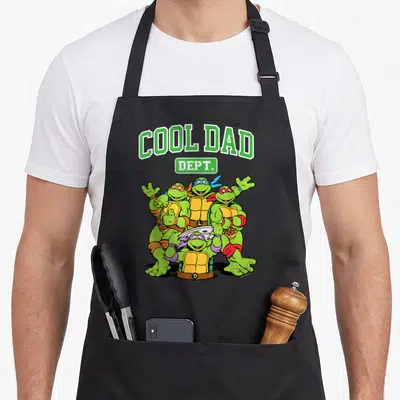 Dept father day apron - COOL DAD DEPT text above four cartoon turtles with blue, red, purple masks and yellow shells — shown on a black cotton canvas apron against a white background