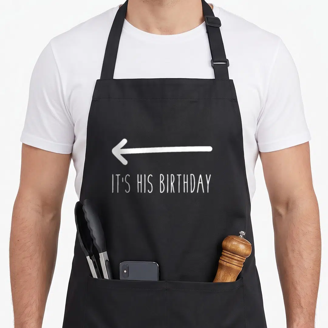 His birthday apron - White arrow pointing left above IT'S HIS BIRTHDAY text on black background — shown on a black cotton canvas apron against a white background