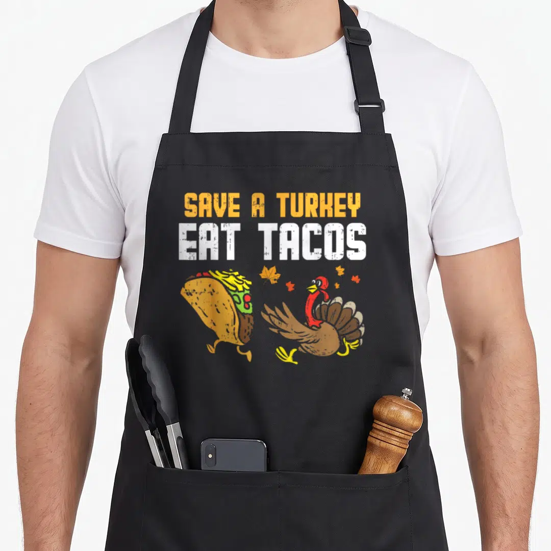 Save thanksgiving day aprons - Running taco and turkey illustration with stacked orange and white distressed text — shown on a black cotton canvas apron against a white background