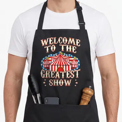 Lion clown aprons - Retro circus tent with arched text reading Welcome to the Greatest Show in red and white stripes — shown on a black cotton canvas apron against a white background