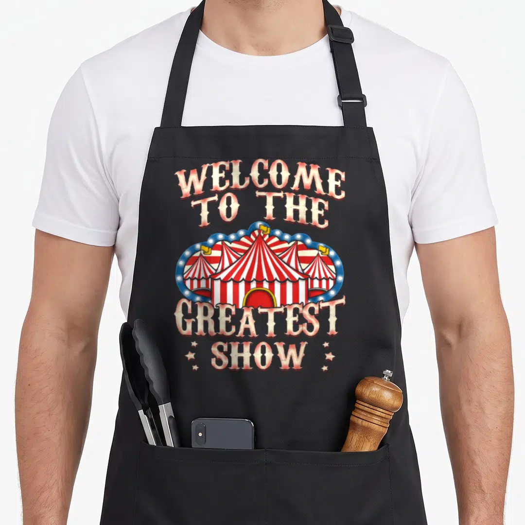 Lion clown aprons - Retro circus tent with arched text reading Welcome to the Greatest Show in red and white stripes — shown on a black cotton canvas apron against a white background
