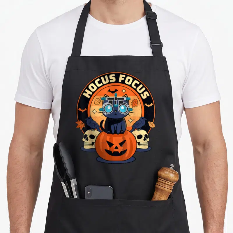 Hocus cat apron - Cat wearing ophthalmic glasses sitting on pumpkin with HOCUS FOCUS text and skulls — shown on a black cotton canvas apron against a white background