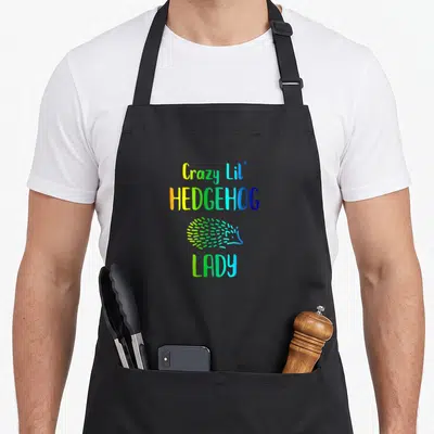 Hedgie rainbow apron - Stacked colorful text reading Crazy Lil' HEDGEHOG LADY with a small hedgehog icon in the center — shown on a black cotton canvas apron against a white background