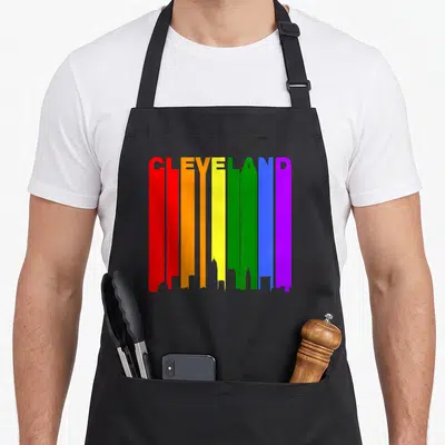 Cleveland gay pride apron - Rainbow vertical bars forming city skyline silhouette with CLEVELAND text at top in red — shown on a black cotton canvas apron against a white background