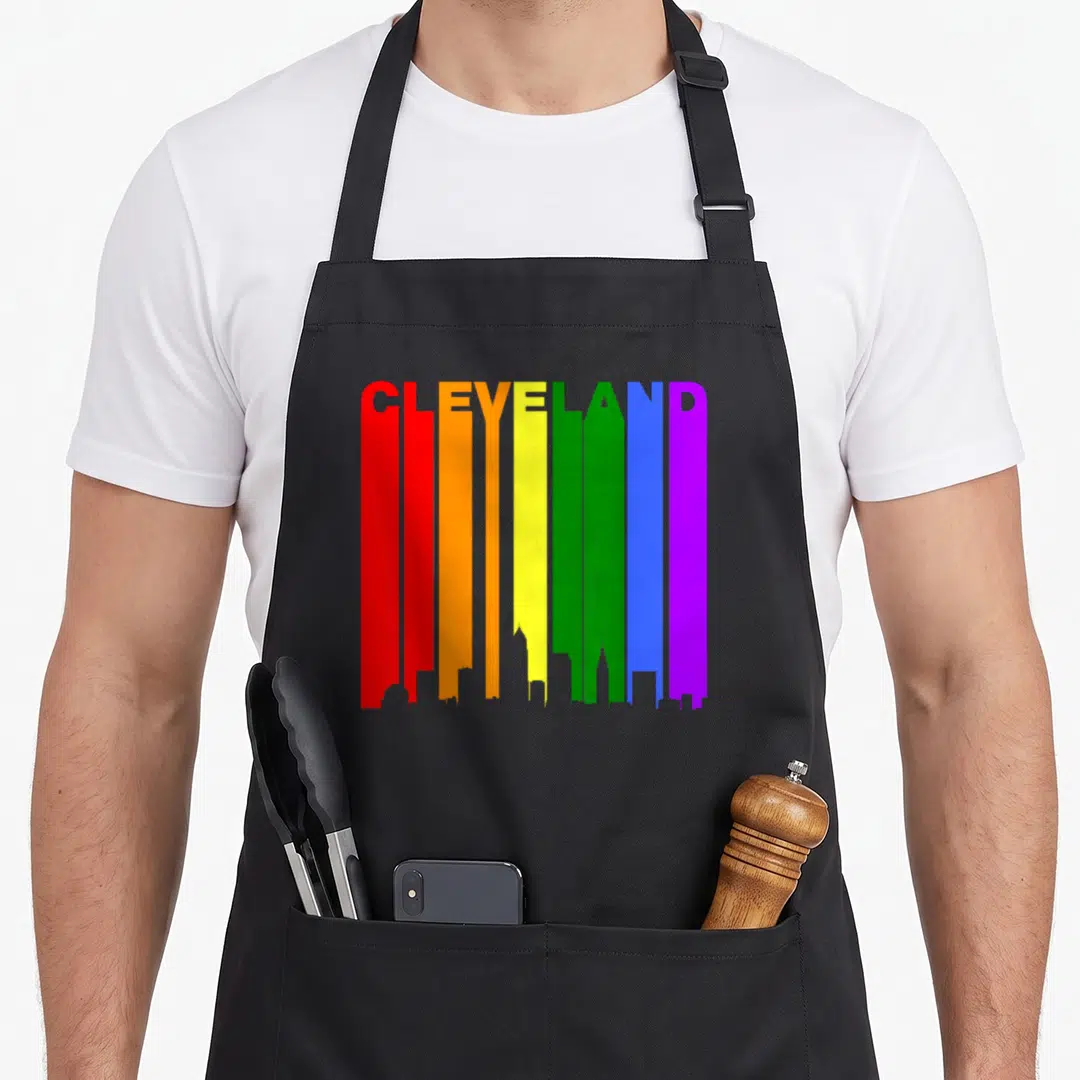 Cleveland gay pride apron - Rainbow vertical bars forming city skyline silhouette with CLEVELAND text at top in red — shown on a black cotton canvas apron against a white background