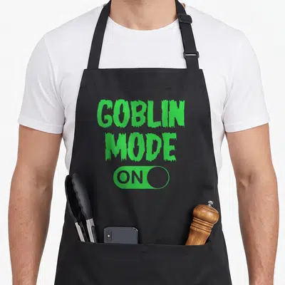 Funny mode goblin apron - Green GOBLIN MODE text above a toggle switch containing the word ON on a grey background — shown on a black cotton canvas apron against a white background