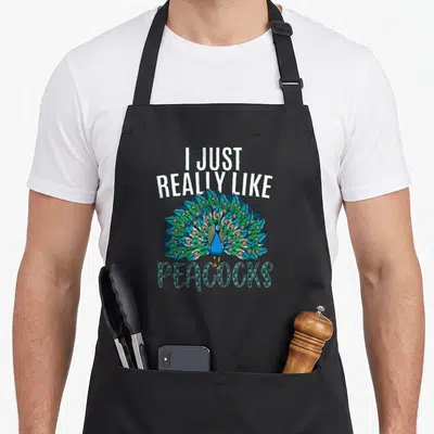 Funny animal bird apron - Peacock with fanned tail in blue green and teal framed by I Just Really Like Peacocks text — shown on a black cotton canvas apron against a white background