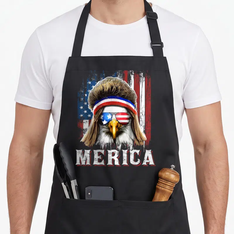 Mullet 4th of july apron - Bald eagle with brown mullet, patriotic sunglasses, and MERICA text on distressed flag — shown on a black cotton canvas apron against a white background