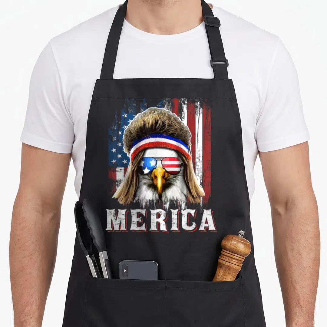 Mullet 4th of july apron - Bald eagle with brown mullet, patriotic sunglasses, and MERICA text on distressed flag — shown on a black cotton canvas apron against a white background