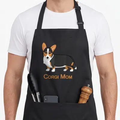 Cardigan dog apron - Cartoon black tan white corgi standing above Corgi Mom text — shown on a black cotton canvas apron against a white background