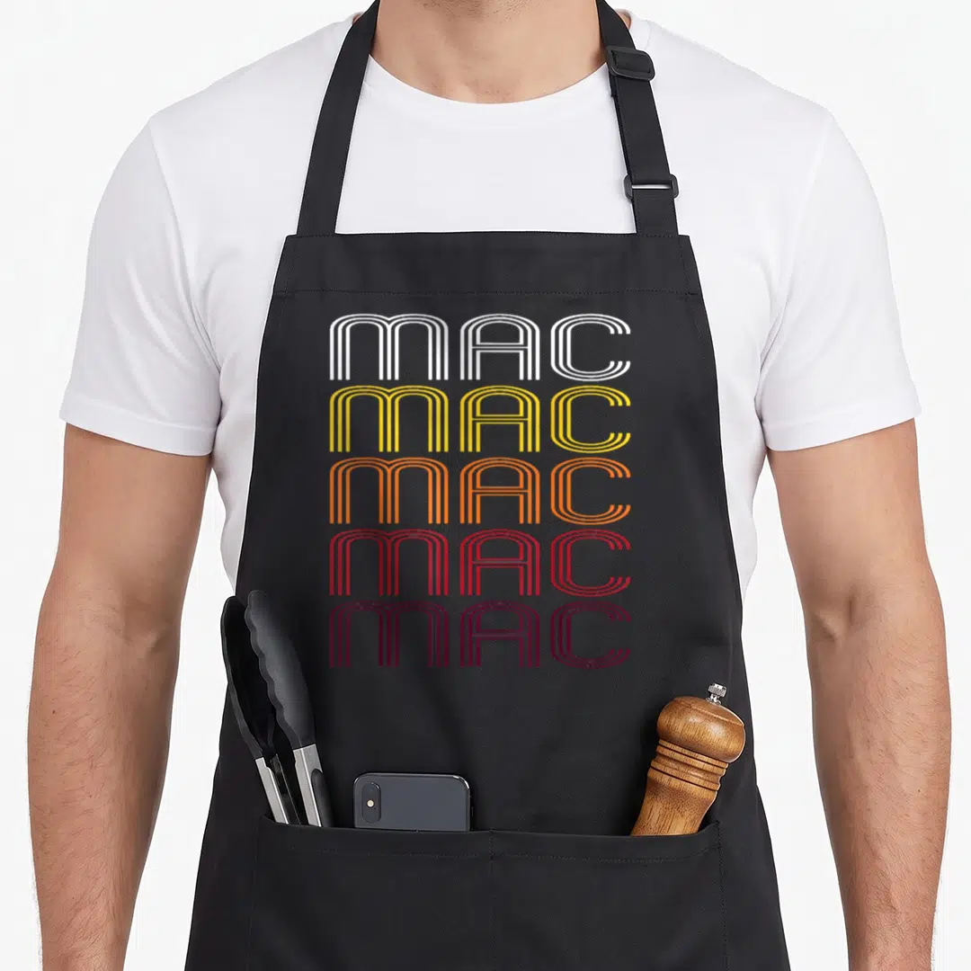 Wordmark mac apron - Five stacked lines of MAC text in white, yellow, orange, red, and maroon retro font — shown on a black cotton canvas apron against a white background