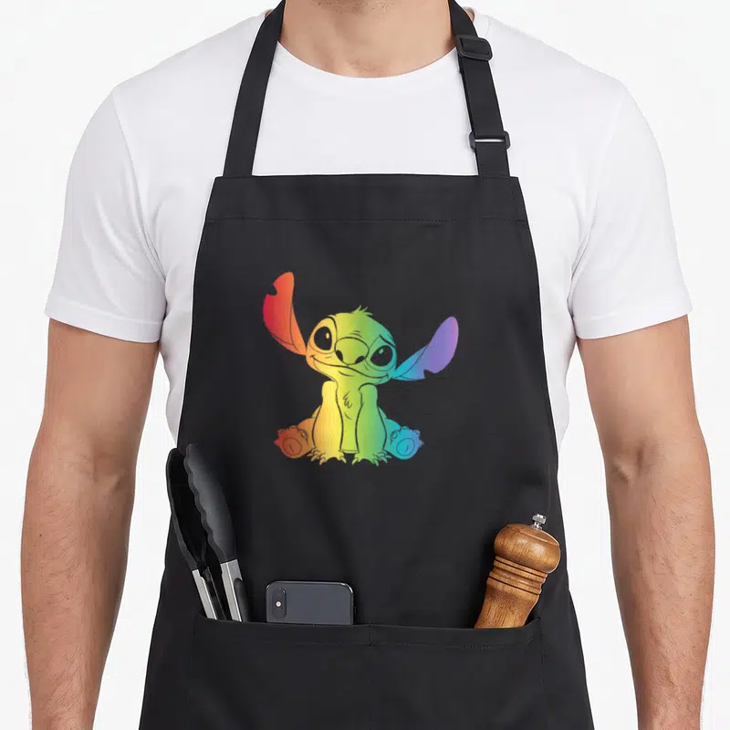 Rainbow stitch apron - Seated cartoon alien figure filled with a rainbow gradient of red, orange, yellow, green, blue — shown on a black cotton canvas apron against a white background