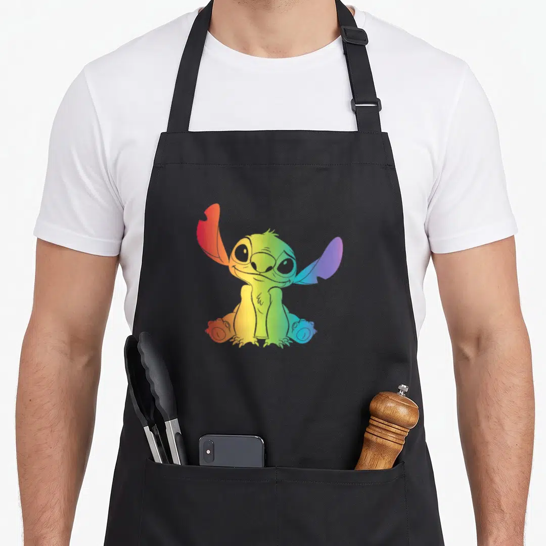 Rainbow stitch apron - Seated cartoon alien figure filled with a rainbow gradient of red, orange, yellow, green, blue — shown on a black cotton canvas apron against a white background