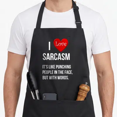 Punching face apron - White I Love Sarcasm text next to red heart with three lines of quote below — shown on a black cotton canvas apron against a white background