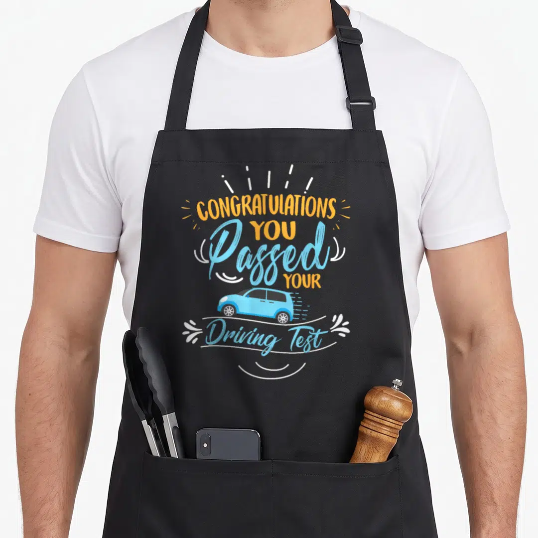 Congratulation driving apron - Orange and blue text 'Passed Your Driving Test' around cartoon car — shown on a black cotton canvas apron against a white background
