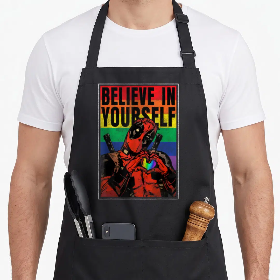 Believe rainbow apron - Deadpool making rainbow heart under stacked BELIEVE IN YOURSELF text on striped background — shown on a black cotton canvas apron against a white background