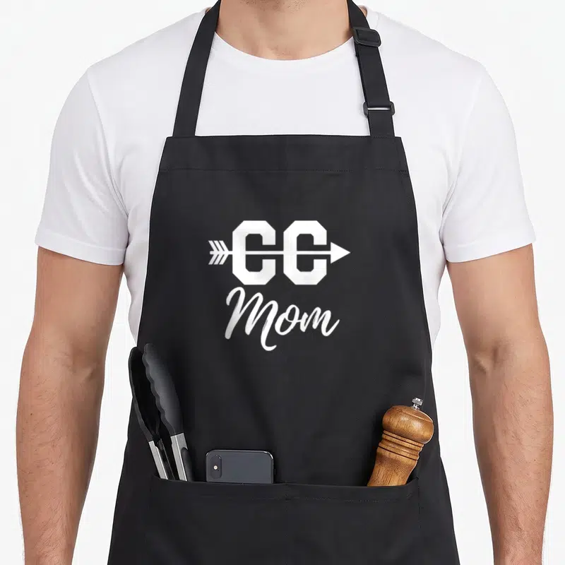 Fan country apron - White CC Mom text with arrow piercing letters stacked above script Mom — shown on a black cotton canvas apron against a white background