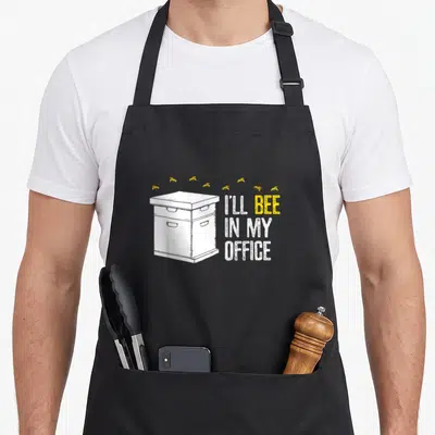 Apiary beekeeper apron - White beehive with yellow bees and I'll Bee In My Office text — shown on a black cotton canvas apron against a white background