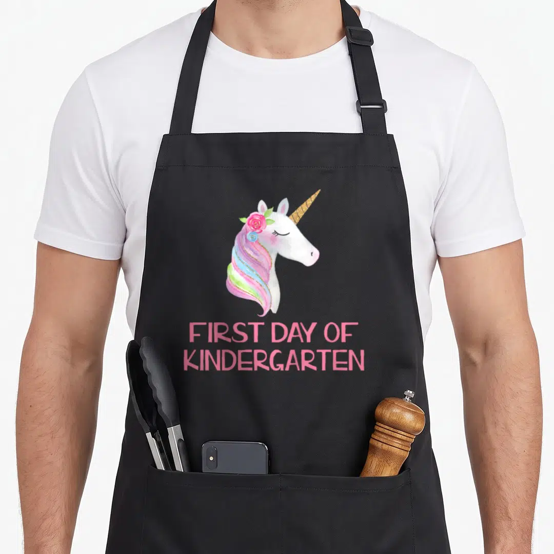 Kindergarten unicorn apron - Watercolor unicorn head with pink mane and flowers above FIRST DAY OF KINDERGARTEN text — shown on a black cotton canvas apron against a white background