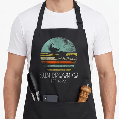Broom salem apron - Grey witch silhouette on teal orange yellow striped circle with SALEM BROOM CO EST 1692 text — shown on a black cotton canvas apron against a white background
