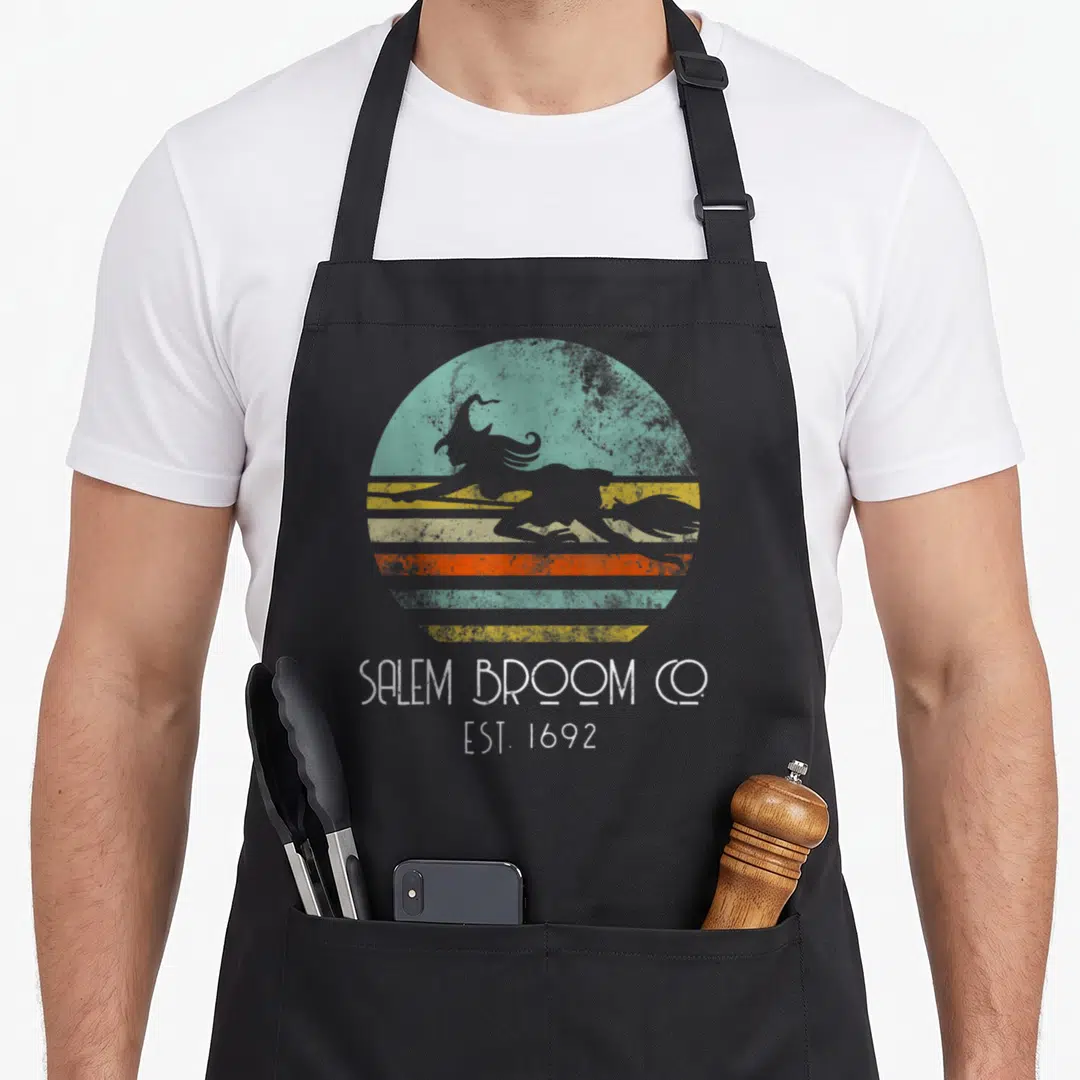 Broom salem apron - Grey witch silhouette on teal orange yellow striped circle with SALEM BROOM CO EST 1692 text — shown on a black cotton canvas apron against a white background