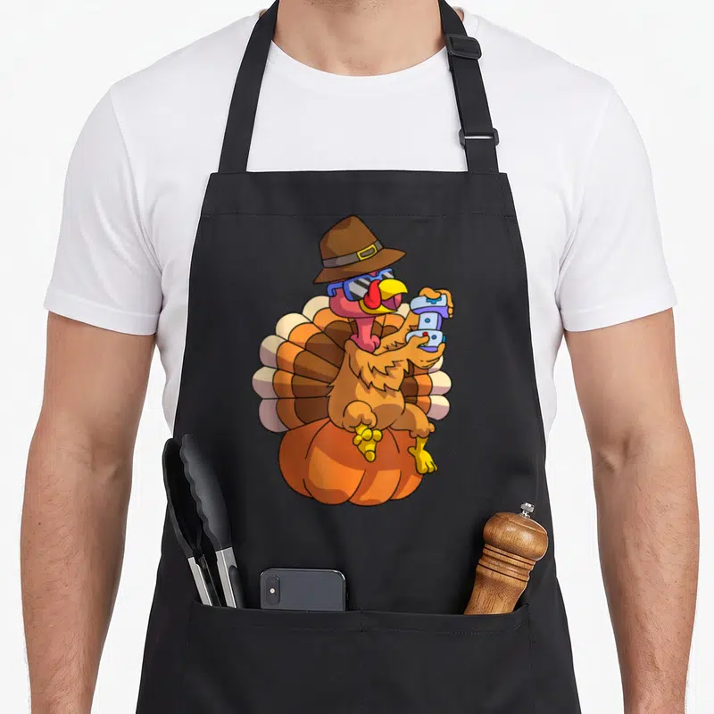 Gamer thanksgiving day aprons - Cartoon turkey with hat sunglasses holding controller on pumpkin in brown orange blue — shown on a black cotton canvas apron against a white background