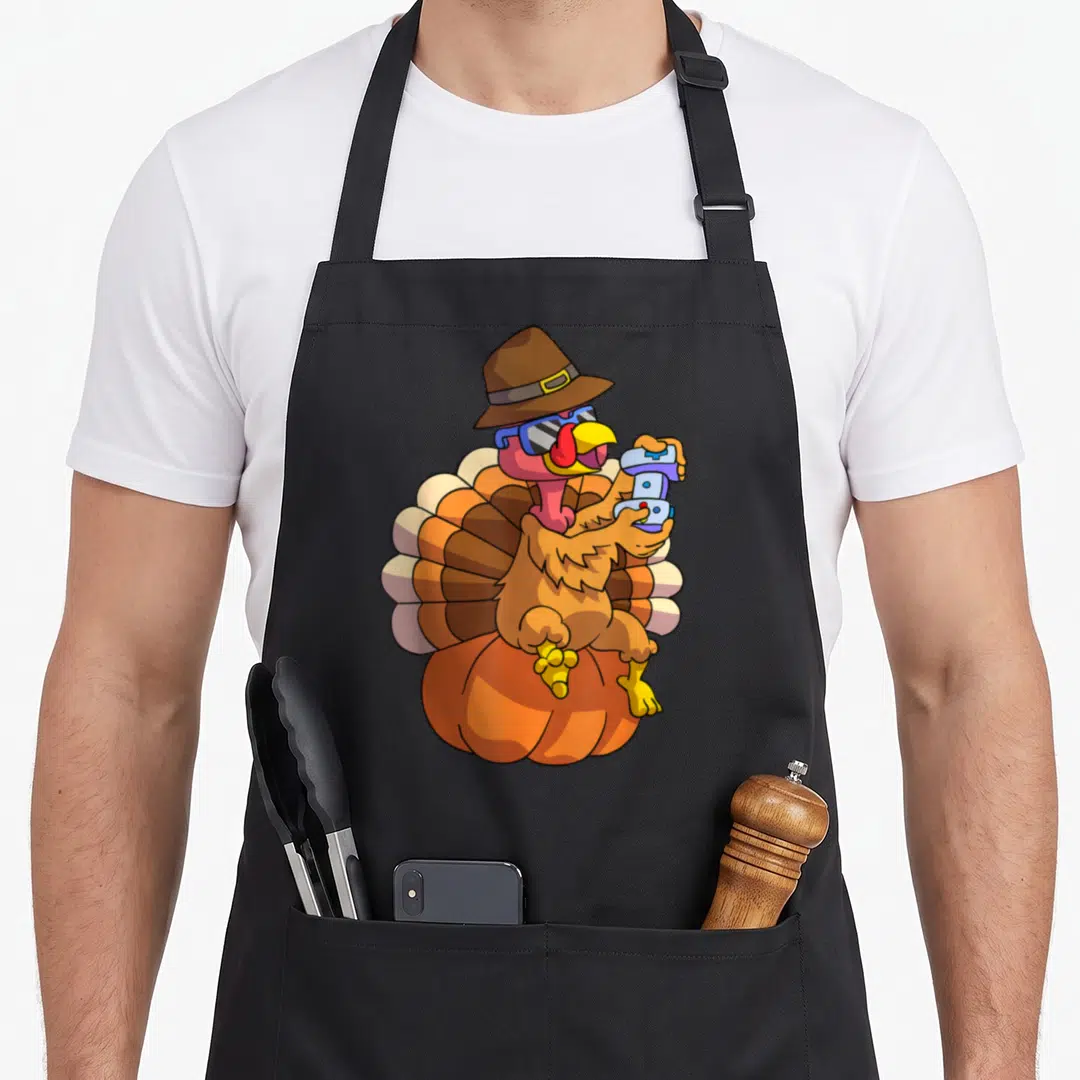 Gamer thanksgiving day aprons - Cartoon turkey with hat sunglasses holding controller on pumpkin in brown orange blue — shown on a black cotton canvas apron against a white background