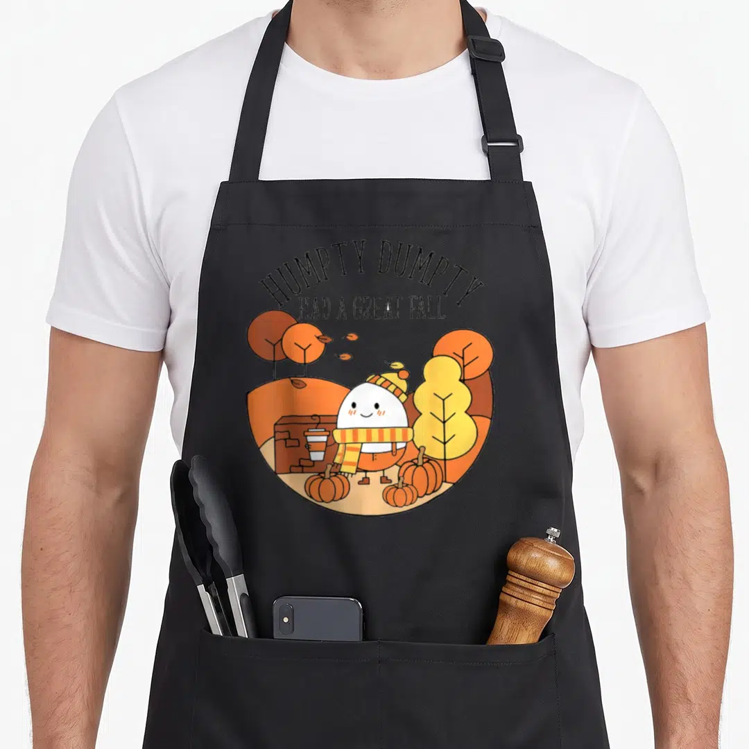 Thankgving cute fall aprons - Cartoon Humpty Dumpty with coffee cup surrounded by pumpkins and autumn trees — shown on a black cotton canvas apron against a white background
