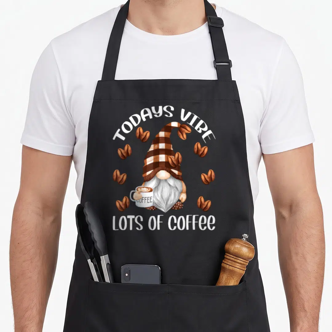 Gnome coffee apron - Watercolor gnome in brown checkered hat holding coffee mug with TODAYS VIBE LOTS OF COFFEE text — shown on a black cotton canvas apron against a white background