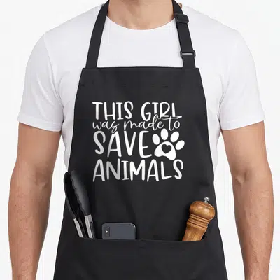 Assistant vet apron - White stacked text This Girl was made to Save Animals next to paw print with heart inside main pad — shown on a black cotton canvas apron against a white background