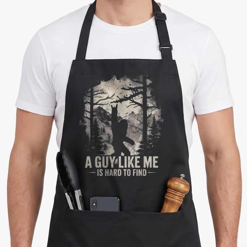 Foot find aprons - Grey cryptid silhouette in beige forest with stacked distressed text below — shown on a black cotton canvas apron against a white background