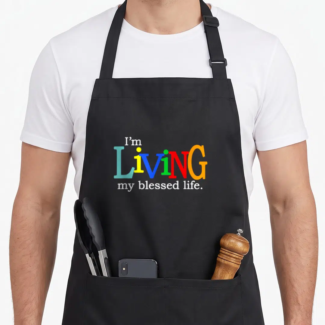 Living christian apron - I'm LIVING my blessed life text in rainbow colors on black background — shown on a black cotton canvas apron against a white background