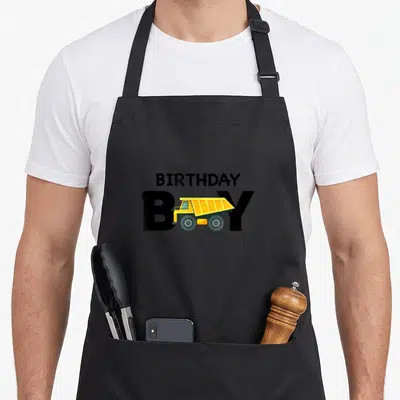 Cute construction truck apron - BIRTHDAY BOY text with yellow dump truck overlapping the O letter — shown on a black cotton canvas apron against a white background