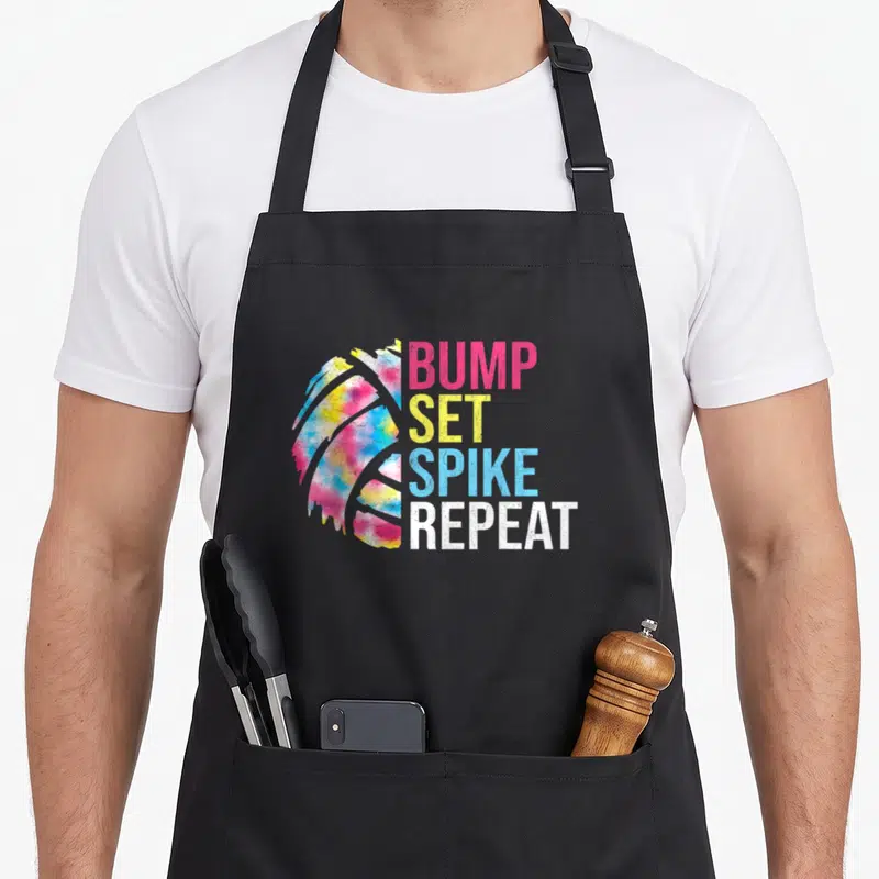Bump volleyball game apron - Tie-dye volleyball graphic next to stacked BUMP SET SPIKE REPEAT text in pink yellow blue — shown on a black cotton canvas apron against a white background