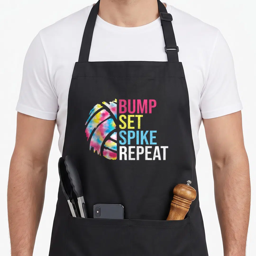 Bump volleyball game apron - Tie-dye volleyball graphic next to stacked BUMP SET SPIKE REPEAT text in pink yellow blue — shown on a black cotton canvas apron against a white background