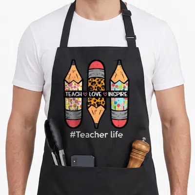Inspire pencil teacher apron - Three cartoon pencils with polka dots and leopard print overlapped by black banner — shown on a black cotton canvas apron against a white background