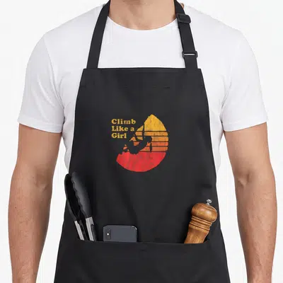 Climbing distressed apron - Climb Like a Girl text beside grey climber silhouette against yellow orange red striped sun — shown on a black cotton canvas apron against a white background
