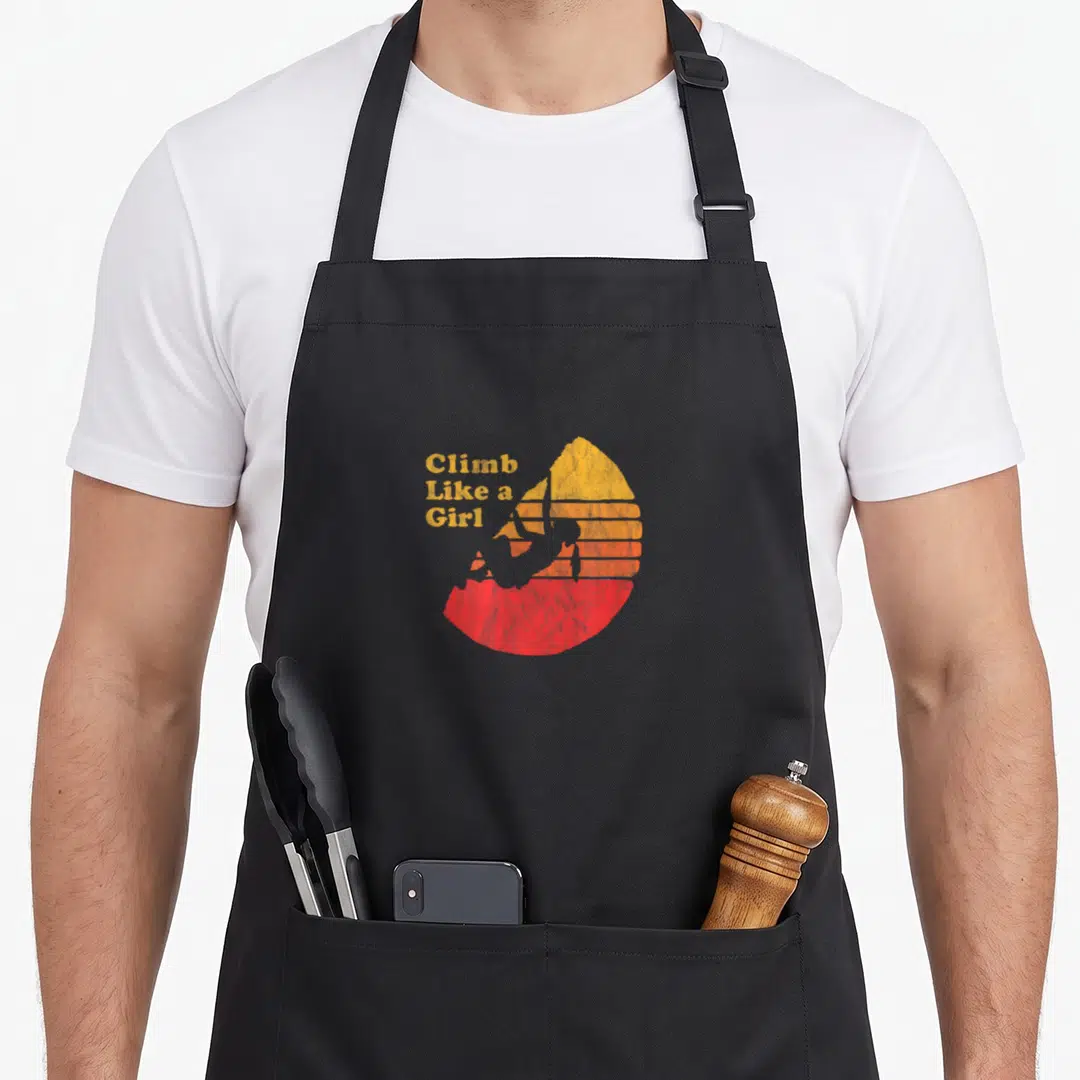 Climbing distressed apron - Climb Like a Girl text beside grey climber silhouette against yellow orange red striped sun — shown on a black cotton canvas apron against a white background