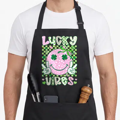Retro lucky st patrick day aprons - Pink smiley face with shamrock eyes and LUCKY VIBES text on checkered background — shown on a black cotton canvas apron against a white background