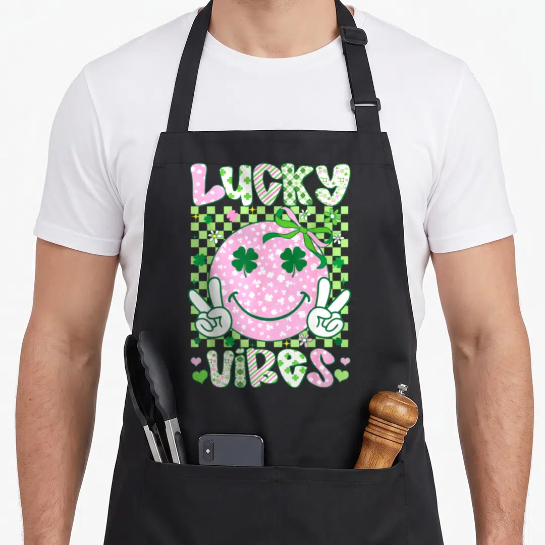 Retro lucky st patrick day aprons - Pink smiley face with shamrock eyes and LUCKY VIBES text on checkered background — shown on a black cotton canvas apron against a white background