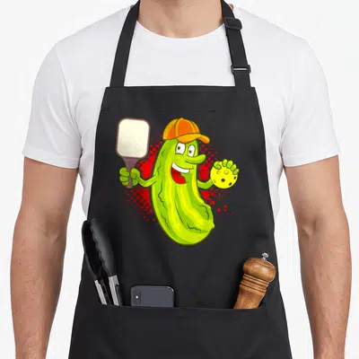 Pickle pickleball apron - Cartoon green pickle wearing orange cap holding paddle and yellow ball on red halftone — shown on a black cotton canvas apron against a white background